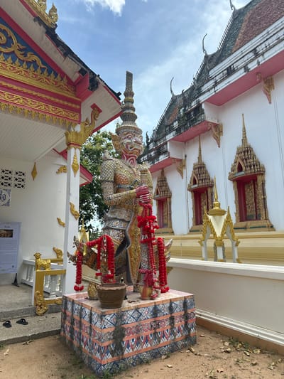 A monumental statue of a mythical giant, likely a Yaksha from Thai folklore, stands guard outside a temple building in Baan Harn, Thailand. The giant is depicted in a dynamic pose, clad in an ornate, multi-colored costume adorned with intricate patterns and gold accents. Red garlands are draped around its neck and arms, suggesting a devotional offering or a ceremonial occasion. The statue is positioned on a decorative pedestal with colorful, repetitive geometric and floral motifs.
Behind the statue, the temple structure features a white facade with prominent red and gold detailing on its roofline and decorative elements. These architectural features, including tiered, pointed roofs with finials and ornate eaves, are characteristic of Thai temple design. The sky above is visible, appearing partly cloudy, suggesting daytime. The ground around the statue is a mix of sand and dry leaves, with some sparse vegetation, indicating an outdoor setting in a warm climate.
Visible text on a white poster board at the base of the statue is in Thai script, though the specific content is illegible in the image. The overall atmosphere is one of reverence and cultural richness, highlighting the artistic and spiritual traditions of the location.