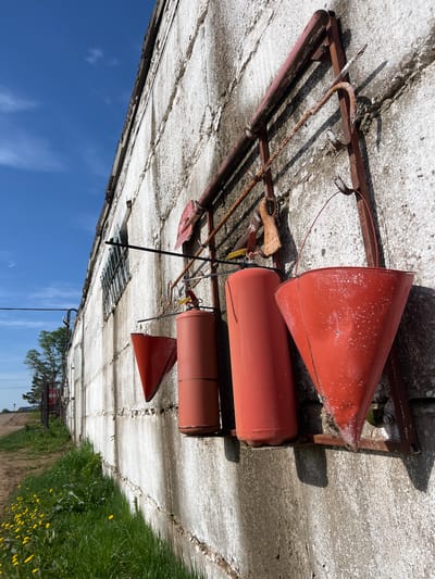 The image depicts a set of antique, red fire extinguishers and associated equipment mounted on a light gray concrete wall, likely an exterior wall. The equipment includes two cylindrical extinguishers, two conical buckets, a small shovel, and a metal rack to hold the equipment. The paint on the extinguishers is weathered and chipped. The metal rack is heavily rusted. The sky is blue. A section of green grass and some dandelions are visible. The location cannot be determined from the image.
