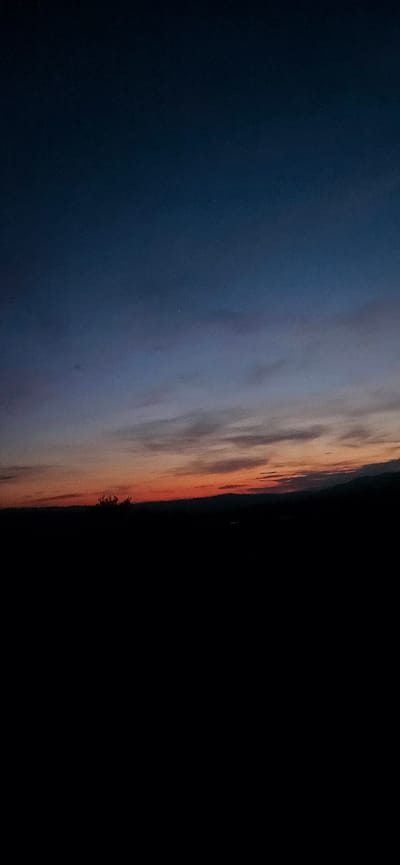 The image depicts a landscape scene at dusk, likely in Momin Prohod, Bulgaria, given the context provided. The sky transitions from a deep blue at the top to shades of orange and red near the horizon, indicating the setting sun. Wispy clouds are visible, catching the last rays of light. The lower half of the image is dominated by a dark, silhouetted landscape, with the faint outlines of hills or mountains in the distance. A few dark, indistinct shapes that could be trees or bushes are present in the foreground on the left. The overall impression is one of twilight settling over a rural or mountainous area. There are no people, animals, or man-made structures clearly visible in the frame.