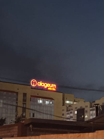 A nighttime exterior shot captures a neon sign on a building, displaying "alageum electric" in bright red lettering. The building itself appears to be an office or commercial structure with large windows. In the foreground, a brick wall and a dark structure partially obscure the view. The sky above is dark and cloudy. The scene is situated in the city of Petropavl, Kazakhstan.