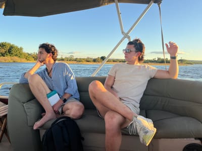 Two men are seated on a boat cruising on a body of water, possibly the Zambezi River near Victoria Falls, Zimbabwe. The man on the left is barefoot, wearing a light blue button-up shirt and khaki shorts, resting his chin on his hand while holding a book. The man on the right is wearing sunglasses, a beige t-shirt, khaki shorts, and dirty white sneakers. He has his right arm raised. A black backpack is on the seat near the barefoot man. The boat is under a canopy structure. In the background, there is vegetation along the shoreline. The sky is clear and bright.