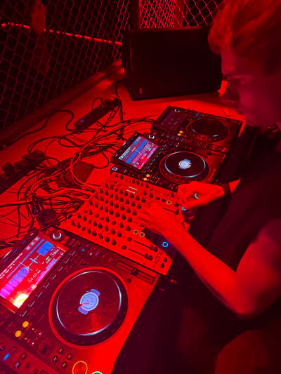 In a vibrant, red-lit nightclub or event space in Shanghai, a DJ is actively performing. The scene is dominated by professional DJ equipment, including two Pioneer DJ CDJ-3000 players and an Allen & Heath Xone:96 mixer. The person, with visible red hair and fair skin, is seen from an overhead angle, with their hands expertly manipulating the controls of the mixer and one of the decks. Their left hand is near the mixer's blue-lit rotary knobs, while their right hand is positioned over the mixer's faders.
The intense red lighting casts a strong, energetic ambiance, with some blue and green highlights emanating from the equipment displays and buttons. The screens of the CDJ-3000s show detailed waveforms, track information, and playback data. On one screen, the track "ANTICORM - Found My Daddy's Gun" is displayed, with a time of "04:40.358" and a tempo of "180.0 Dm". Another screen shows "05:59 180.0 Dm" and "00071 DID".
Behind the equipment, a tangled mass of cables is visible, typical of a professional setup. A black speaker with the brand "ARIE" is positioned to the left. A metal mesh structure, possibly part of a railing or stage, is visible in the lower left, adding to the industrial, club-like atmosphere. The focus of the image is on the DJ's interaction with the equipment, highlighting the technical and performative aspects of DJing.
