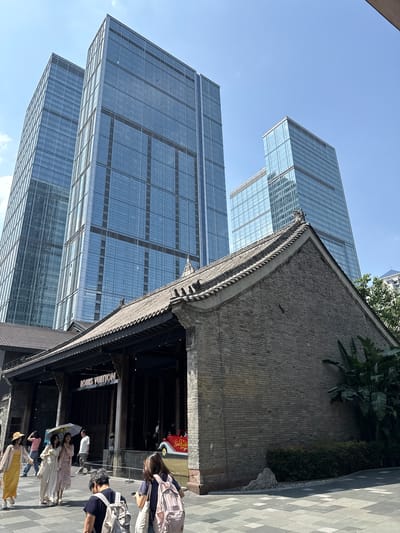 The image captures a street scene in Jinjiang District, China, showcasing a juxtaposition of architectural styles. In the background, modern skyscrapers with glass facades rise against a clear blue sky. In the foreground, a traditional Chinese building with a dark-tiled roof and intricate wooden detailing stands. People, dressed in a variety of modern clothing, walk along the paved street. The entrance to a "LOUIS VUITTON" store is visible, blending the traditional facade with a modern luxury brand. The overall impression is a blend of old and new, indicative of Jinjiang District's urban landscape.