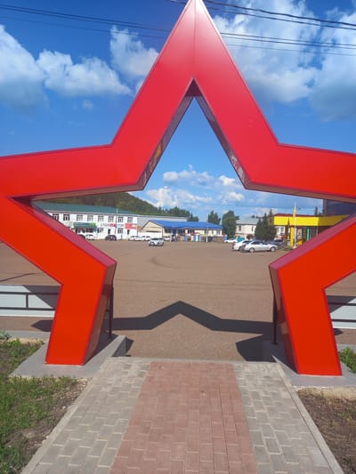 The image shows a large, bright red five-pointed star sculpture framed in the foreground. The star is viewed from an angle that makes it appear hollow, with the sky and a town visible through its opening. The background features a gravel-covered area, several buildings with different colored roofs (white, green, blue, and yellow), parked cars, and a distant treeline with a hill. The sky is a vibrant blue with fluffy white clouds. The ground in the immediate foreground is paved with interlocking bricks in a pattern of red and gray. There are no people visible in the image. The location is identified as the town square in Sterlibashevo, Russia, indicated by the text "СТОЛ И СТУЛ" (Table and Chair) visible on one of the buildings, a common business name in Russia.