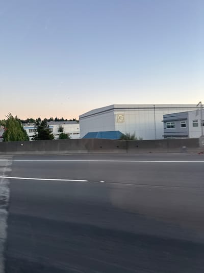 This is a photograph of a commercial building in Burnaby, Canada, as seen from a roadway. The building is a large, light-colored structure with a prominent "G" logo on its facade. To the left of the main building, there is another structure that appears to be an office building with windows. The buildings are situated behind a dark concrete barrier, and a highway or major road is visible in the foreground, with a white line marking a lane. The sky above is clear and pale blue, indicating it is either early morning or late afternoon. Trees and some smaller buildings are visible to the left of the main structure. There are no people or vehicles clearly identifiable in the image, and the scene appears to be static. The overall impression is one of an industrial or commercial area within a suburban or urban setting.