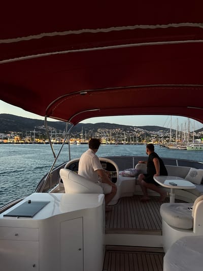The image shows two men on a boat, with one steering and the other sitting nearby. The boat appears to be docked or moving slowly in a harbor with many sailboats visible in the background. The scene is set against a backdrop of a town built on a hillside, illuminated by lights, suggesting it is dusk or early evening. The sky is a soft blue, indicating a clear evening. The water is calm, reflecting the lights from the town and the sky.
The man at the helm is wearing a white shirt and is focused on steering. The other man, wearing a black t-shirt, is seated slightly behind and to the side, looking towards the town. Both men appear relaxed. The boat itself is modern, with a wooden deck and white seating areas. A red canopy extends over part of the deck. The overall mood is peaceful and serene, with the gentle rocking of the boat and the beautiful view of the illuminated town. The location is identified as Bodrum, Turkey, a popular coastal town known for its marinas and ancient architecture. No visible text is present in the image.