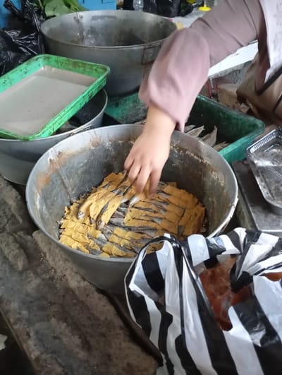A person, wearing a brown long-sleeved shirt, is arranging dried fish in a silver bucket in a market. The fish are laid out in a grid pattern and appear to be packaged in thin sheets of yellow paper. The fish are being arranged on top of other dried fish in the bucket. There is a black and white plastic bag next to the bucket and a green plastic container to the left. The scene is located in a market in Kutawaringin, Indonesia. The coordinates for this location are -6.953 latitude and 107.524 longitude. The environment is cluttered and appears to be outdoors.