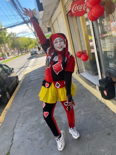 A woman in a theatrical costume with makeup is standing on a sidewalk holding a microphone. The costume is predominantly red, black, and yellow, featuring diamond, spade, and heart motifs. She wears white sneakers and a head covering. A Coca-Cola sign and red balloons are visible in the background next to a window, likely indicating a business or shop. A portable speaker sits on the sidewalk nearby. A street scene with cars, trees, and buildings is visible, and based on the urban setting with distinctive architecture and the wire infrastructure for power lines, this scene appears to be located in Quito, Ecuador.