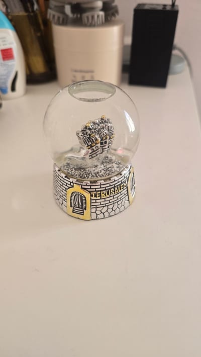 A snow globe sits on a white surface. The snow globe is clear glass and contains a miniature replica of the Dome of the Rock, which is silver with gold accents. The base of the snow globe is designed to resemble a stone wall with a gold archway and the word "JERUSALEM" written in gold lettering. In the background, out of focus, are various objects including a bottle of lotion, a beige cylindrical container, and a black electronic device. The setting appears to be an indoor surface, possibly a desk or table.