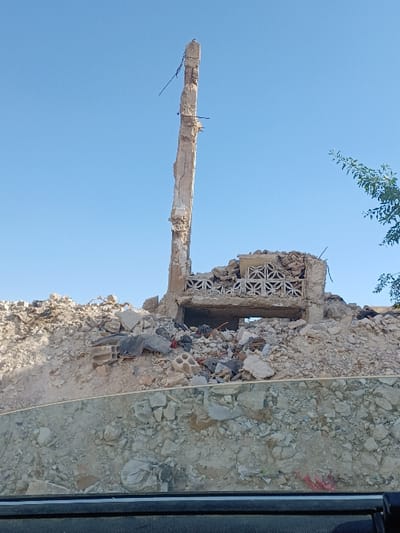 The image depicts a scene of extensive structural damage in Al-Qadam Municipality, Syria. The central focus is a severely compromised reinforced concrete structure against a clear, bright blue sky. A tall, slender, and heavily damaged concrete pillar, with exposed rebar, stands prominently on the left side, appearing to be a remnant of a larger building. To its right and slightly lower, a section of a collapsed structure is visible, featuring a horizontal concrete element adorned with repeating geometric patterns, possibly a decorative balustrade or window screen. An opening, indicative of a former window or doorway, is present beneath this decorative section.

The ground surrounding these remnants is covered by a large mound of debris and rubble, composed of broken concrete, masonry, and other construction materials. In the foreground, the image includes the upper section of a vehicle's dashboard and a reflective glass surface, through which the scene is viewed. A small cluster of green leaves from a tree or bush is visible on the far right against the sky. No individuals or active subjects are present in the frame. The environment is characterized by destruction and the aftermath of a significant impact or demolition event.