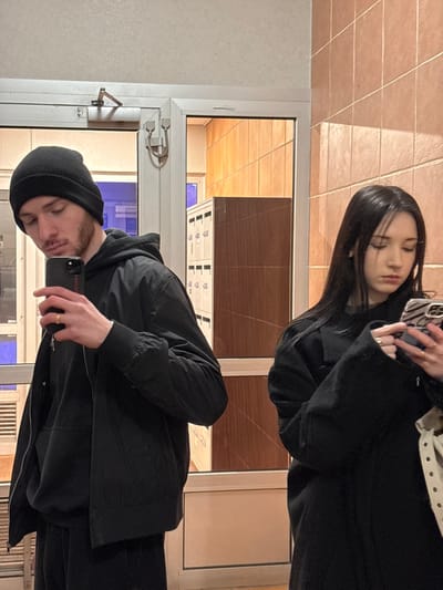 A mirror selfie shows a man and woman. The man wears a black beanie and jacket, holding a phone to take the picture. He has a gold ring on his finger. The woman stands behind him, also in black, holding a phone with a zebra-striped case. The background is a tiled floor and wall with a white-framed window. The reflection shows a brown wall inside.