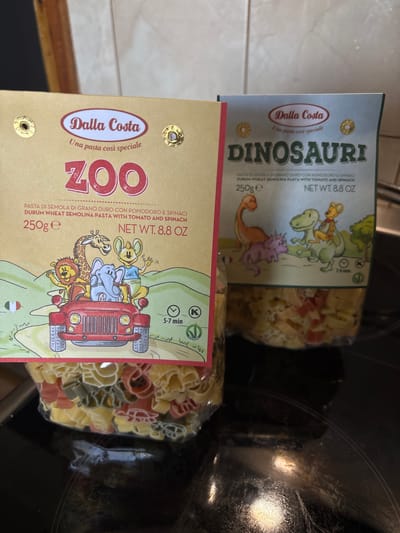 Two bags of Dalla Costa pasta are displayed on a black stovetop. The bag on the left is yellow and features the word "ZOO" in large red letters. It depicts cartoon animals on a red jeep, with a giraffe, lion, and elephant visible. The bag on the right is light blue and has the word "DINOSAURI" in green letters. This bag shows cartoon dinosaurs. Both bags contain pasta shaped like animals and dinosaurs. The pasta in the bags is a mix of yellow, red, and green colors. The background shows a blurred kitchen environment. The location of Cheshnegirovo, Bulgaria, is not discernible from the image.