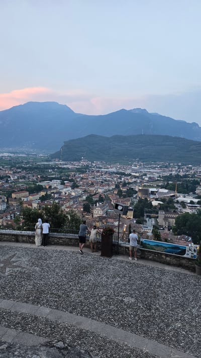 The image depicts a scenic overlook of a town nestled between mountains. The viewpoint is from a stone-paved area with a low metal railing. Several people are visible, standing and observing the panorama. One person in the foreground is taking a photo with their phone. Further back, two people are seated, and another two are standing and looking out. The town below is densely packed with buildings, exhibiting a mix of architectural styles and terracotta roofs. Lush green trees are interspersed throughout the urban landscape, and the imposing presence of dark, tree-covered mountains forms a dramatic backdrop.

The time of day appears to be late afternoon or early evening, as indicated by the soft, diffused light and the sky, which shows a hint of pink and orange hues near the horizon, suggesting twilight. The weather seems pleasant, with clear skies over the mountains and a gentle haze or light cloud cover. The location is identified as Riva del Garda, Italy, which aligns with the mountainous terrain and the style of the town.

Notable details include a planter with flowers near the railing and a lamppost. A framed picture or informational display is mounted on a wall behind the railing. The overall atmosphere is one of peaceful observation and appreciation of the stunning natural and urban landscape. There is no visible text in the image.