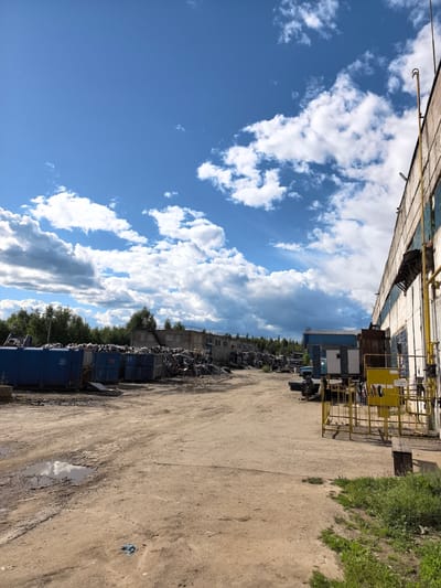 This is a wide, outdoor shot of a salvage yard or industrial facility on a sunny day with scattered clouds. The scene depicts a lot of accumulated material, including large blue dumpsters, piles of what appears to be scrap metal or refuse, and various buildings in the background. A dirt road runs through the foreground, with some puddles and tire tracks visible. To the right, a large, light-colored industrial building dominates the frame, with yellow pipes and conduits running along its exterior. A yellow metal fence encloses an area with some equipment. There are no people clearly visible in the image. The weather appears to be fair, with a bright blue sky. The overall impression is one of a working industrial or recycling site. Some text is visible on the dumpsters, including numbers like "30" and "51," and Russian text on a yellow sign within the fenced area. The location context is Ivanovo, Russia.