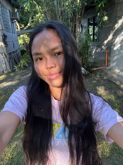 This image, captured in Carmen, Philippines, depicts a young female taking a close-up outdoor selfie. She possesses long, straight, dark hair and dark eyes, looking directly at the camera with a slight smile. A distinct line of sunlight and shadow traverses her face diagonally from the upper left forehead to the lower right chin, with the right side of her face (viewer's left) brightly illuminated and the left side in shadow. She is wearing a light lavender short-sleeved t-shirt featuring a partial yellow and blue geometric design on the front, along with a thin gold-colored necklace. Her arms are partially visible on the lower left and right edges of the frame, indicating she is holding the camera.

The background consists of a semi-rural or rural outdoor environment under bright sunlight. To the left, a weathered wooden structure, appearing to be a simple building or hut constructed from vertical wooden planks, is visible. Behind it, dense green foliage and trees are present. To the right, a light grey concrete block wall with a rectangular dark-framed window is situated. A dense cluster of bamboo plants with slender culms and green leaves stands behind the concrete wall. An orange PVC pipe is attached horizontally and then vertically along the concrete wall. The ground beneath is a mix of dirt, sparse green grass, and small stones, exhibiting pronounced shadows consistent with direct sunlight.
