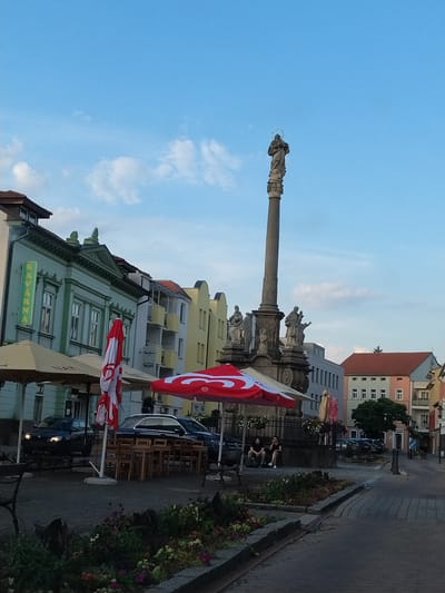 The image shows a view of the Strakonice square in Strakonice, Czechia, during daylight. The square features a prominent Marian column with statues at its base and a statue of the Virgin Mary at the top. Several buildings with distinct architectural styles line the square, including a pastel green building with the word "Kavarna" visible and a multi-story yellow building. Umbrellas, some red and white, are set up, suggesting an outdoor seating area. Tables and chairs are visible beneath the umbrellas. Two dark-colored cars are parked on the street. In the foreground, there is a flower bed with various plants. Two individuals are seated on a bench near the base of the Marian column. The sky is blue with scattered clouds.