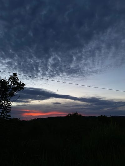 The image captures a sky at dusk in a location consistent with the climate of Sosail, Indonesia. Dark, ominous clouds cover the left side of the sky, while the right side displays a gradient of colors, from grey and light blue to streaks of orange and red near the horizon. A dark treeline marks the edge of the land, silhouetted against the twilight sky. A power line stretches across the image. A silhouetted tree with visible leaves appears in the lower portion of the image.