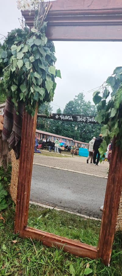 The image shows an outdoor scene at what appears to be a cultural event or festival in the village of Старые Быги, Russia. A large, rustic wooden frame, possibly part of an art installation or entrance arch, is in the foreground, adorned with leafy branches and some white flowers. Through the frame, a paved area with people milling about is visible. In the background, a row of colorful banners or flags hangs across stalls or market booths. A wooden sign with white lettering is suspended above the stalls. People are dressed casually, and some are carrying umbrellas, suggesting possibly overcast or drizzly weather. The environment is green, with grass and trees visible.