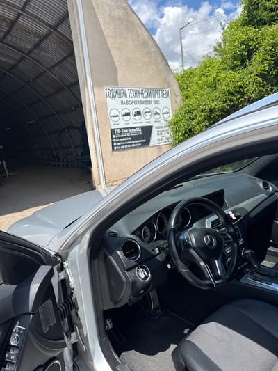 The image shows the interior and exterior of a silver Mercedes-Benz vehicle parked in front of a building with a large arched entrance. A sign on the building's exterior wall advertises "ГОДИШНИ ТЕХНИЧЕСКИ ПРЕГЛЕДИ НА ВСИЧКИ ВИДОВЕ ППС," which translates to "Annual Technical Inspections for All Types of Vehicles." Below this, the sign indicates the company name as "ГПР - Елин Пелин ООД," a "Technical Inspection Center - Elin Pelin Ltd." and provides contact information including an address at "гр. Елин Пелин, бул. „ЯнкоСинчец“ 119," which is Elin Pelin, Yanko Sintets Blvd. 119. It also lists working hours: Monday to Friday from 08:00 to 17:30 and Saturday from 09:00 to 13:00. The environment appears to be a service or garage area, with the interior of the garage visible through the entrance, showing what looks like inspection equipment. Lush green foliage is visible behind the building on the right side of the image, and the sky above is partly cloudy. The car's interior, featuring a black leather steering wheel with Mercedes-Benz branding, dashboard, and fabric seats, is prominently displayed. The driver's side door is open. A person in dark clothing is partially visible inside the garage entrance. The overall scene suggests the vehicle is at a vehicle inspection station in the town of Elin Pelin, Bulgaria.