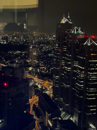 An elevated night-time view of the Shinjuku skyscraper district in Tokyo, Japan, is captured. The image features a dense urban landscape illuminated by a multitude of building lights and streetlights. In the foreground, several modern skyscrapers dominate the scene. One prominent building on the right displays a distinctive architectural design with triangular, pyramid-like structures at its apex, illuminated by white and green lights. The sides of this skyscraper are lined with numerous lit windows, suggesting occupancy. Red warning lights are visible on the upper levels of various buildings. Below the skyscrapers, a complex network of elevated highways curves through the city, with car headlights and taillights creating streaks of light on the roadways. In the background, the urban sprawl extends to the horizon, a vast expanse of smaller buildings with scattered lights, fading into the dark, hazy night sky. Reflections of interior lights and possibly a ceiling fixture are visible in the foreground window through which the photo was taken, slightly obscuring the view of the cityscape.