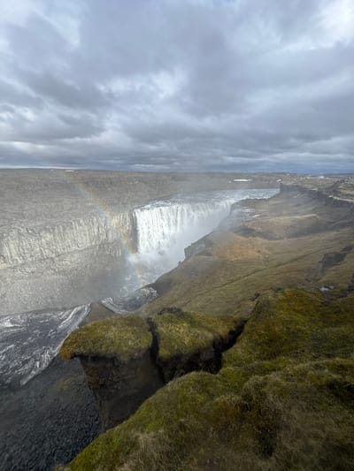 The image captures a powerful waterfall cascading over a rugged, moss-covered cliff face under a cloudy sky. The main body of water plunges dramatically, creating a significant amount of white mist that rises from its base.

To the left of the waterfall's main drop, a faint, ethereal rainbow is visible within the water spray, suggesting sunlight is interacting with the mist. The cliffs flanking the waterfall are steep and appear to be composed of dark, possibly volcanic rock, which is extensively covered in lush, vibrant green and brownish-green moss and some sparse, dry grasses. These textured, mossy slopes are particularly prominent on the right side of the frame, showing the rugged contours of the terrain.

The sky is largely overcast, filled with various shades of grey and white clouds, contributing to the dramatic and somewhat subdued lighting of the scene. No people or visible text are present in the image. The natural grandeur, geological features, and vegetation are consistent with landscapes found in regions like Iceland.