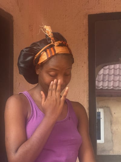 A woman reclines with her eyes closed, her right hand partially covering her face. She wears a purple tank top and a black satin bonnet with a patterned orange and brown headscarf tied around the edge. The background features a textured wall with a dark-framed window above. The angle of the shot is slightly tilted, giving a sense of informality.