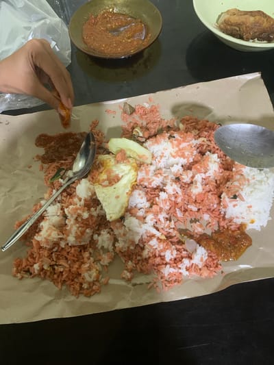 The image captures a close-up view of a meal served on brown paper, likely in Sungai Meriam, Indonesia. The meal consists of rice that is partially pink and partially white, mixed with small pieces of vegetables and what appears to be fried egg. A spoon is placed on top of the rice. To the left, a hand is visible, holding a piece of food and appearing to be in the process of eating. There are also two small bowls in the background, one containing a reddish sauce and another with a piece of fried food. The scene is set on a dark, reflective surface, possibly a table.