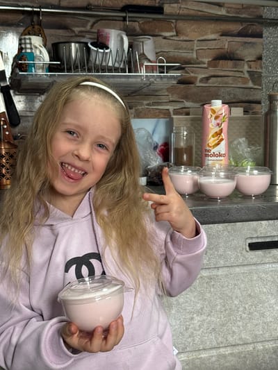 A Caucasian girl with long blonde hair and wearing a lavender sweatshirt with a black logo is holding a sealed cup of pink dessert and pointing at similar desserts in a cupboard. The background includes a carton of milk and kitchenware. The location is in Kurganinsk, Russia.