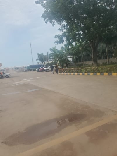 The image captures a scene in Gwagwada, Nigeria, depicting a paved area with several vehicles. In the foreground, the light-colored asphalt shows patches of dampness, suggesting recent rain. A yellow line runs across the pavement, demarcating a parking or traffic lane.
Further back, multiple cars are parked, including a white SUV and a red sedan. Beyond the cars, a fleet of trucks is visible, some of which appear to be cargo trucks with tarpaulin covers. A tall, slender tower, possibly a communication mast, rises in the background against a hazy sky.
The environment is characterized by greenery, with palm trees and other large trees lining the background, interspersed with buildings that appear to be part of a commercial or industrial complex. Two individuals are walking in the mid-ground, facing away from the camera, dressed in casual attire. One is wearing dark clothing, and the other, a lighter brown jacket and pants. Another person, wearing an orange vest, is standing near a white car on the left side of the frame, seemingly engaged in an activity. The overall impression is that of a logistics hub or a large parking area within a business district.