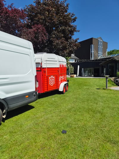 The image depicts a white van connected to a small red and white trailer parked on a green lawn. The trailer has "www.letzkola.lu" printed on its side. In the background, there's a modern, dark-colored house with large windows and an adjacent tree with reddish-brown leaves. The sky is clear and blue. The photograph appears to be taken during daytime with bright sunlight. The location is Lenningen, Luxembourg, indicated by the website printed on the trailer.