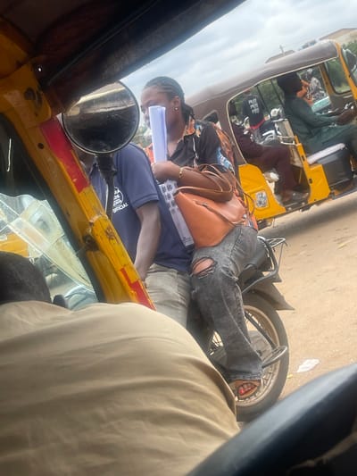 The image shows a group of people inside and around a yellow auto-rickshaw, a common mode of transport in Nigeria. The scene appears to be set in a village, given the "Galadimawa Village, Nigeria" location context. The lighting suggests it's daytime, and the slight overexposure of the sky indicates bright, possibly sunny weather.
One person, a young woman, is seated, holding a white rolled-up document or paper. She is wearing ripped denim jeans and a dark t-shirt with a graphic design. A tan-colored shoulder bag rests on her lap. Her expression is difficult to discern clearly, but she appears focused on the document.
Other individuals are visible within the auto-rickshaw, including a person in a dark green garment and another in a maroon shirt. The interior of the auto-rickshaw is somewhat cluttered with bags and other items. The exterior of the vehicle is a vibrant yellow with some red accents. The road beneath the auto-rickshaw appears to be unpaved or dirt.
A side mirror is attached to the front of the auto-rickshaw, reflecting parts of the scene. The overall impression is one of everyday life and movement within a Nigerian village setting. The presence of the document being held suggests potential activity related to travel, work, or education.