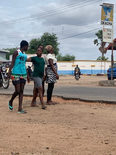 The image captures a street scene in Shendam, Nigeria, under an overcast sky. Three women are walking from left to right on a reddish-brown dirt shoulder alongside a paved road. The woman on the left wears a light blue sleeveless top with dark Chinese characters, dark patterned shorts, and light blue flip-flops. The central woman wears a dark green short-sleeved top, grey shorts, and light brown flip-flops, with her left arm extended towards the first woman's back. The woman on the right wears a white top with puff sleeves, a black and white striped skirt, dark leggings, and dark sandals, with a light brown head covering.
In the mid-ground to the right, a fuel station sign displays "OKACHA" at the top with a blue and yellow emblem featuring a white bird/hand. Below, another section lists "PETROL," "DIESEL," and "KERO" with associated prices, including "840 PETROL" and "650 KERO." Behind the sign, a palm tree and a partially visible blue car are present.
Further in the background, a low white wall with a blue base stripe features multiple red "NO SMOKING" inscriptions. A man on a motorcycle, wearing a dark green shirt, is seen riding in front of this wall. To the far left, another motorcycle is parked. The immediate foreground consists of a rough, stony dirt surface.
