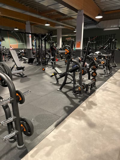 An interior view of a commercial fitness center in Aubange, Belgium, is depicted. The primary training area features dark grey rubber matting, transitioning to a lighter beige tiled floor on the right, separated by a black ribbed floor strip. The ceiling is high, exposing metallic ductwork, light-colored wooden beams, and multiple white-shaded pendant lights. Walls are predominantly dark green/grey, with visible text "FREE WEIGHT" on the back wall and a partially visible sign stating "BETTER TODAY THAN YESTERDAY." The left wall displays orange and white curved markings, suggestive of a sports court boundary.
Numerous pieces of black-framed strength training equipment are present, many featuring orange accents on weight plates, pin selectors, or adjustment points. In the immediate foreground, a black weight plate rack with several silver barbells and orange-rimmed black plates is visible on the left. To the right of the foreground, a black plate-loaded machine is equipped with orange weight plates. A black adjustable utility bench is positioned mid-ground on the left, near a multi-station cable machine. Another similar bench is visible further right.
Several individuals are present within the gym. One dark-skinned person wearing a black shirt and black pants stands near the cable machine in the mid-ground left, appearing to observe or adjust the equipment. Another individual, wearing a dark top, is partially visible bent over near the plate-loaded machine in the mid-ground right. A person in a pink top is seated on a bench in the far background on the left. Additional blurry figures are sparsely distributed throughout the background, engaged in various fitness activities or standing near equipment. Shadows from the equipment and individuals are cast across the floor, particularly distinct on the lighter tiled section.