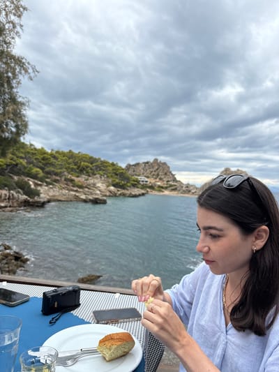 The image captures an adult female seated at an outdoor table beside a body of water in the Municipal Unit of Loutraki - Perachora, Greece, under an overcast sky. The woman, with dark brown hair and black sunglasses resting on her head, is wearing a light blue collared shirt, a gold-colored chain necklace, and small gold hoop earrings. She has light pink fingernail polish and wears rings on her left index finger, right ring finger, and right index finger. Her attention is focused on her hands as she tears a small piece of a yellow object, likely lemon.
On the table, covered with a blue and white checkered tablecloth and a solid blue section, a white ceramic plate holds a half-eaten bread roll, a silver fork, and a knife. To the left, a clear glass contains water, and a smaller clear glass with liquid is also visible. Two dark-cased smartphones and a black compact film camera with a strap are positioned on the table.
The background features a calm, dark blue-green sea. The coastline on the left is characterized by densely vegetated hills and rocky outcrops. A tall, slender tree with green needles is partially visible in the upper left. On the right, the coastline continues with more rocky formations, scattered vegetation, and a distant white building with a red roof on a hillside. The sky is uniformly covered with layered grey clouds. A wooden edge or railing is visible in the immediate foreground, indicating an elevated dining area overlooking the sea.