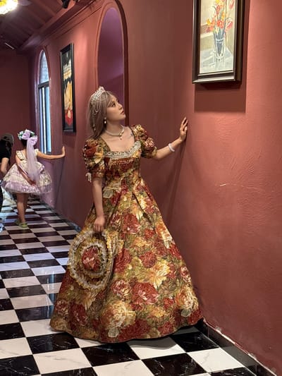 The image shows a woman wearing a floral gown and holding a matching hat. She is posing in a room with a checkerboard floor and paintings on the wall. The walls are a reddish-brown color. In the background, another woman in a lilac gown is visible. The room appears to be part of a building. The location is in Baqiao, China.