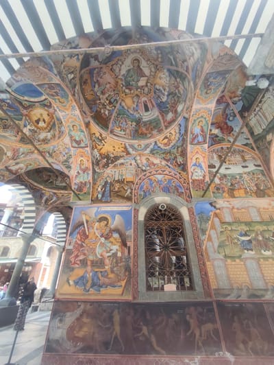 The image depicts the exterior frescoes of the Rila Monastery in Rilski manastir, Bulgaria. The elaborate murals cover the arched ceiling and walls of an open-air structure, featuring numerous religious scenes and figures, primarily rendered in vibrant colors. In the foreground, a large fresco shows an angel battling a winged beast. Below this, a frieze of human figures in various poses is visible. A barred window is set into the wall. To the left, a person is standing and appears to be taking a photograph, holding a phone up to their eye. The architectural elements of the monastery, including columns and striped awnings, are also visible. The overall scene is rich in detail and artistic expression, showcasing a significant religious and cultural site.