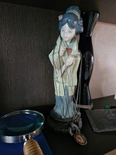 This image depicts a still life on a shelf, likely in a domestic setting in Bremen, Germany. The central subject is a porcelain figurine of a geisha, adorned with several necklaces. To the left of the figurine, a magnifying glass with a wooden handle rests on a blue surface. Behind the figurine, a tall, black, vase-like object stands. The background consists of a dark wooden paneling on the left and a textured, light-colored surface on the right. The objects are arranged on a dark shelf. The lighting suggests an indoor environment, possibly natural light coming from the side or overhead.