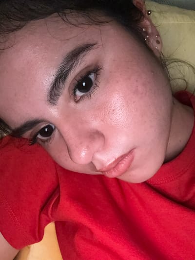 A close-up, rotated image shows a person lying on a yellow surface, possibly a pillow or cushion. The person is wearing a red garment, likely a shirt or top. Their face is prominent, displaying features such as dark eyes with thick eyelashes, dark eyebrows, a nose with visible pores, and slightly glossy lips. The skin shows some texture and minor imperfections. The upper part of an ear with multiple piercings is visible in the top left corner. The background is blurred, further emphasizing the person's face. No specific location in Austin, Texas, can be determined from the visual information provided.