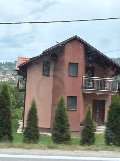 The image shows a brown house with a dark tiled roof, situated in a hilly, green landscape. There are several evergreen trees planted in front of the house, creating a natural fence. The house has multiple windows, some dark and rectangular, and a balcony with a white metal railing on the upper floor. The sky appears overcast, suggesting a cloudy day. A thin wire runs vertically through the left side of the frame, possibly a power line or telephone wire. In the background, more houses and trees are visible on the slopes of the hills, indicating a rural or semi-rural setting. The overall impression is of a residential area in a temperate climate. There is no visible text in the image.