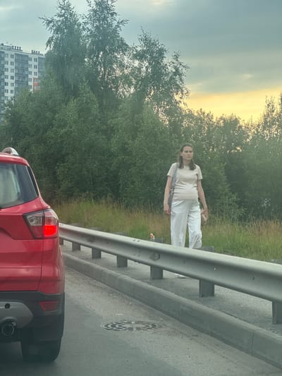 A pregnant woman stands on the side of a road in Novoe Devyatkino, Russia, near a grey metal guardrail. She is wearing a light-colored t-shirt and loose white pants, with a bag slung over her shoulder. In the foreground, the rear of a red SUV is visible, its brake light illuminated. The background consists of a dense line of green trees and a modern multi-story building, with a hazy yellow and grey sky above. The ground is paved with asphalt and gravel, with a concrete curb and a manhole cover visible. The overall scene appears to be an urban or suburban setting during the day, possibly late afternoon given the lighting conditions.