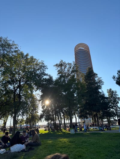 The landscape features a towering cylindrical building reaching into a clear blue sky. Trees dot the foreground, with people gathered beneath their shade on a grassy area. Sunlight filters through the leaves, creating bright flares. The scene suggests a public gathering in an urban park setting with a modern skyscraper as a focal point.