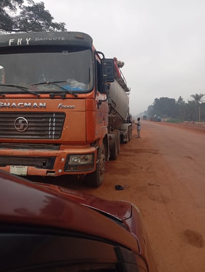 In Imilike, Nigeria, a cement truck, identified as a SHACMAN F2000 with "FKY DANGOTE" branding on its upper front, is parked on a dusty, reddish-brown road. The truck's windshield is cracked. A second cement truck and several other vehicles are visible further down the road, suggesting a potential traffic or a roadside stop. A single individual, a young male wearing a light-colored shirt and dark pants, stands near the cement truck, looking at a mobile phone. The environment appears to be rural, with trees and vegetation bordering the road. The sky is overcast, indicating a cloudy day. The foreground is occupied by the hood of a dark red vehicle, partially obscuring the scene.