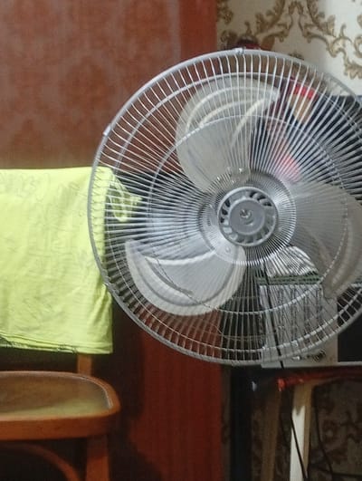 The image depicts a close-up view of a fan positioned in a room. The fan is the primary subject, a metallic oscillating fan with silver blades and a protective grill. Its blades are partially visible, suggesting it may be in operation or poised to be. The background is partially obscured, but a portion of a wooden chair with a yellow cloth draped over it is visible to the left. The wall behind the fan appears to have patterned wallpaper. The lighting suggests an indoor setting, possibly during the daytime, but no specific time can be determined. There are no people visible in the image. The overall scene is static and domestic. The location context is Termez, Uzbekistan, but there are no explicit visual cues in the image to confirm this specific location. No text is visible in the image.