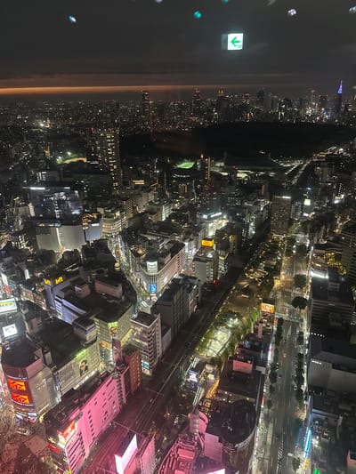 An aerial view captures the vibrant city of Shibuya, Japan, at night. The urban landscape is dominated by brightly lit buildings, illuminated streets, and distinct structures. The Tokyo Plaza can be identified by its unique cylindrical design and illuminated signage. Pedestrians and cars are visible on the busy streets, adding to the dynamic atmosphere. An exit sign is visible, as well as city lights extending to the horizon.