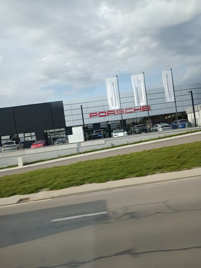 The image shows a modern Porsche dealership located in Pancharevo, Bulgaria. The building has a sleek, contemporary design with a dark corrugated metal facade and a white section featuring the prominent red "PORSCHE" logo. Three white banners with the Porsche logo are displayed outside the building. In front of the dealership, several Porsche cars are visible through the large windows, including a red sports car, a silver SUV, a silver coupe, a white sedan, and a blue sedan. The dealership is situated next to a road, with a green grassy area separating it from the asphalt. The sky above is cloudy, with a mix of grey and white clouds. The image appears to have been taken from a moving vehicle due to motion blur.