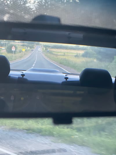 The image is taken from inside a vehicle, looking forward through the rearview mirror and the rear windshield. The perspective suggests the camera is positioned behind the front passenger seat. The scene captured is a rural road in Niva, Russia.

In the foreground, the dark interior of the car is visible, including the backs of headrests and parts of the dashboard. The rearview mirror is prominently featured, reflecting the road ahead.

The road itself is paved and appears to be a two-lane highway with a white dashed line marking the center. It curves gently to the right as it recedes into the distance. Various road signs are visible along the roadside, including a yellow triangular sign with a black symbol and a yellow rectangular sign.

On either side of the road, there are grassy fields and trees, indicative of a rural or semi-rural environment. In the distance, there are a few vehicles on the road, including what appears to be a light-colored car or SUV with its headlights on. The sky is overcast or hazy, with the sun diffused, creating a soft light. The overall impression is of a journey through the Russian countryside.