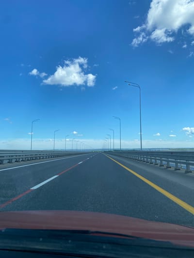 The image, correctly oriented horizontally, depicts a vast, open highway or bridge stretching into the distant horizon under a bright blue sky. The perspective is from inside a vehicle, with a reddish-brown section of the car's interior (likely dashboard or door) visible on the far right.

The road surface is dark asphalt, marked with a prominent solid yellow line on the left and a dashed white line bordering a continuous red line on the right side of the visible lane. Along the left edge of the road, a long, grey metal guardrail runs parallel to the asphalt, extending far into the distance. Beyond the guardrail, a seemingly endless line of tall, slender streetlights, each with a double-arm fixture, recedes sharply towards the hazy horizon.

The sky is a dominant feature, a brilliant, clear blue with several distinct, fluffy white cumulus clouds scattered across it, primarily on the left side. The horizon line where the sky meets distant land or water is very low and hazy, emphasizing the expansive, open nature of the location.

The scene captures a moment of travel on a sunny, clear day, likely midday, conveying a sense of journey and openness. There are no people or other vehicles visible. The characteristics strongly suggest a long bridge or causeway, aligning with the provided location context of Soroch'i Gory, Russia, known for its bridge over the Kama River. There is no visible text in the image.