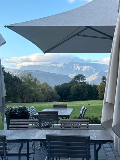 The image shows a view through a patio setting towards distant mountains. The patio is furnished with wooden tables and chairs, partially shaded by patio umbrellas. A green lawn is visible beyond the patio, with trees and shrubbery leading to the base of the mountains. The mountains in the background are rugged, with some snow visible. The sky is partly cloudy. The image may be taken in Stellenbosch Nu, South Africa.