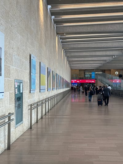 The image depicts a long, covered walkway within what appears to be an airport or a large public transportation hub in Tzafria, Israel. The walkway is lined with a textured, light-colored stone wall on the left, featuring several framed exhibits or posters. A prominent "FIRE" sign is visible on a metallic door on the left wall. On the right side of the walkway, a polished wooden floor reflects the overhead lighting and the activity within the space. A metal handrail runs along the length of the walkway.
In the distance, a crowd of people is seen walking, many with luggage, indicating travel. Digital signs displaying advertisements and information are visible in the background, including a "JR/DUTY FREE" sign and a clock. The ceiling is characterized by a modern design with exposed structural beams and integrated lighting. The overall atmosphere is one of transit and a busy public space.