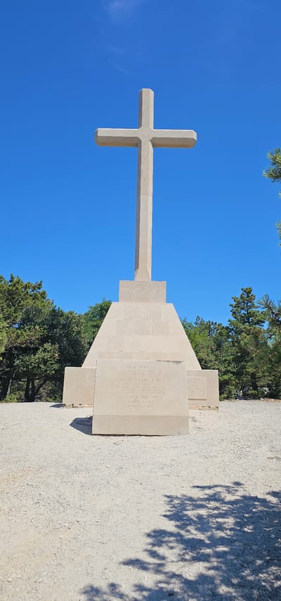 The image shows a tall, stone cross monument situated on a gravel-covered area, likely an outdoor memorial or religious site. The cross is made of light-colored stone and stands against a clear, bright blue sky. The base of the cross is a multi-tiered stone structure with an inscription on a prominent plaque on the front. The surrounding environment includes lush green trees and some visible pine branches, suggesting a natural, possibly hilly or mountainous location. The overall impression is one of solemnity and remembrance. This monument is located in the town of Općina Bol, Croatia, on the island of Brač.